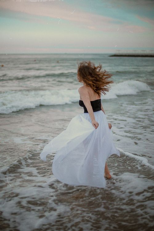 Free A woman in a white dress dances freely on a beach, embracing the sea waves. Stock Photo