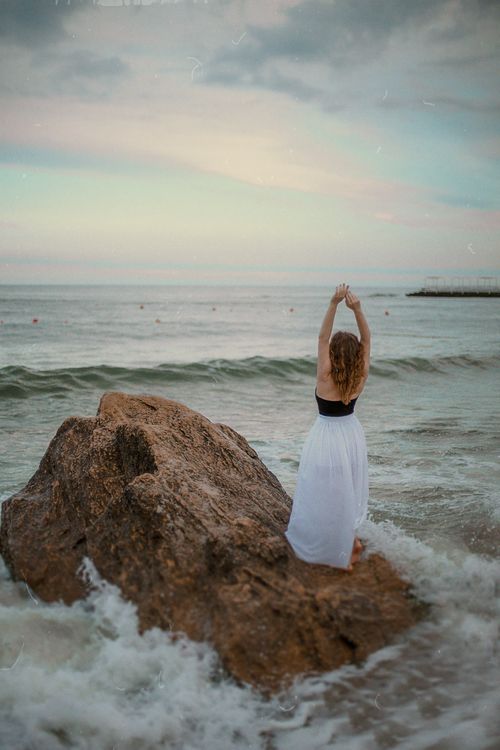 Free A woman in a white dress stands on a rock by the ocean, arms raised against a vibrant sunset sky. Stock Photo