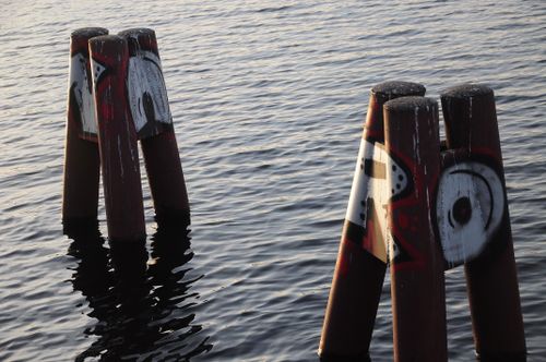 Free Artistic graffiti adorns weathered wooden posts protruding from calm water, creating a striking visual. Stock Photo