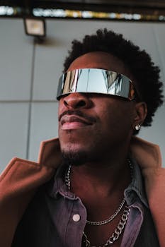 A fashionable man with reflective sunglasses, chains, and a jacket in an urban setting.