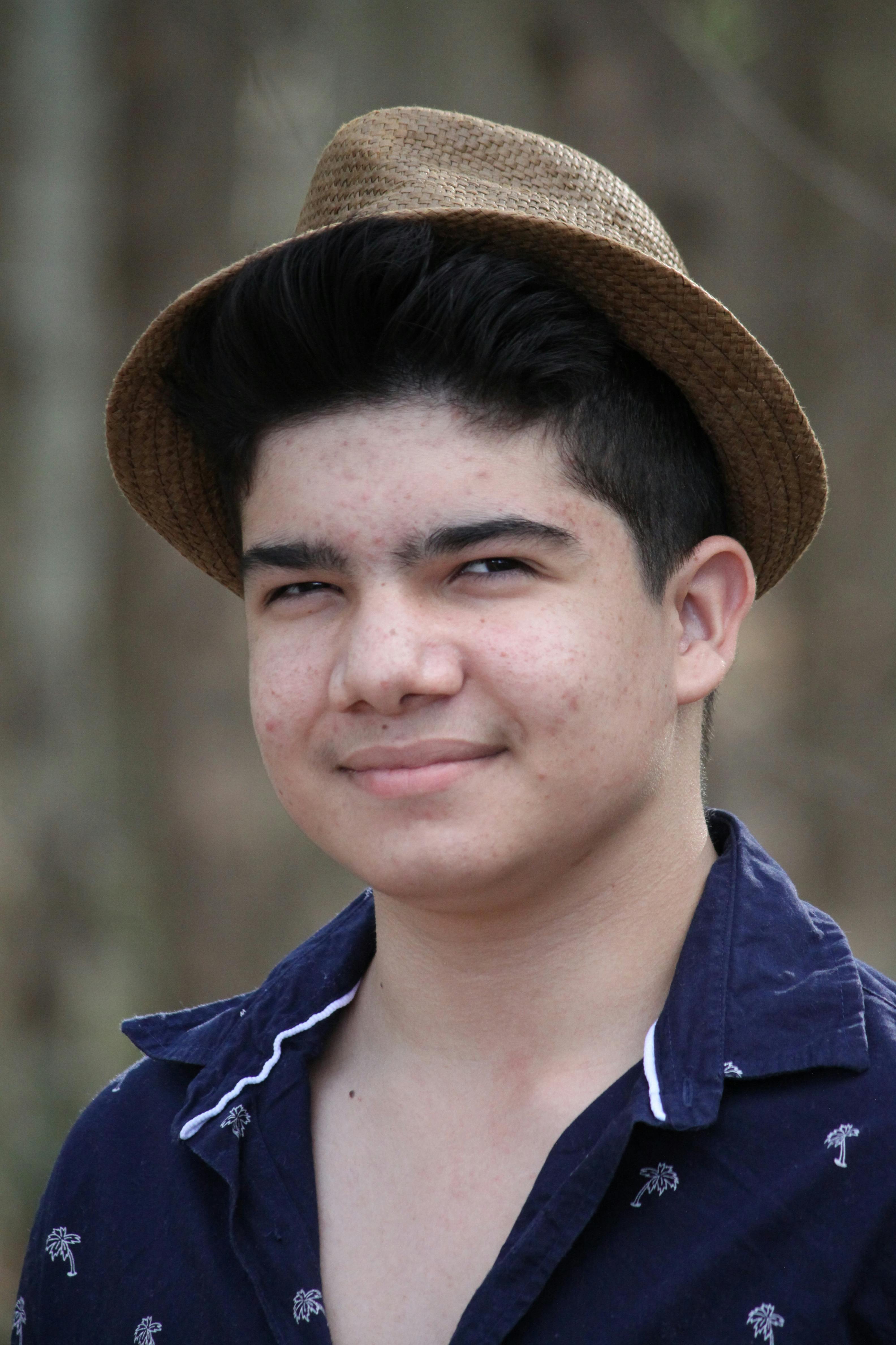 Portrait of a young man wearing a fedora hat, smiling outdoors against a blurred background.