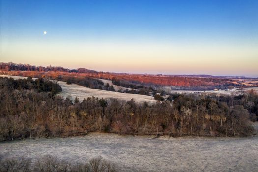 Aerial view of a serene winter landscape with tree lines and moon in blue sky at dawn in Minnesota.