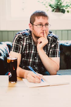 一个戴着眼镜的年轻人在咖啡馆里边思考边写作，手边放着一杯饮料