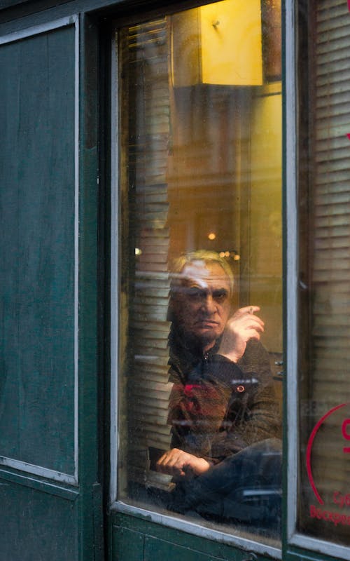 Man in Black and Brown Jacket Sitting Beside Glass Window