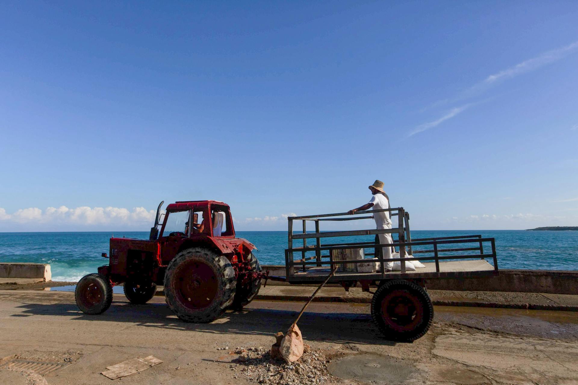 A red tractor and trailer by the sea under a clear blue sky.