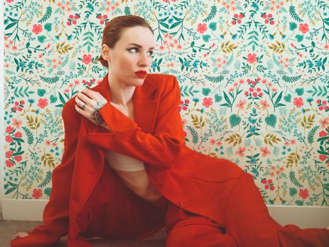 Elegant woman in a red blazer sits against a vibrant floral wallpaper, exuding style and confidence.