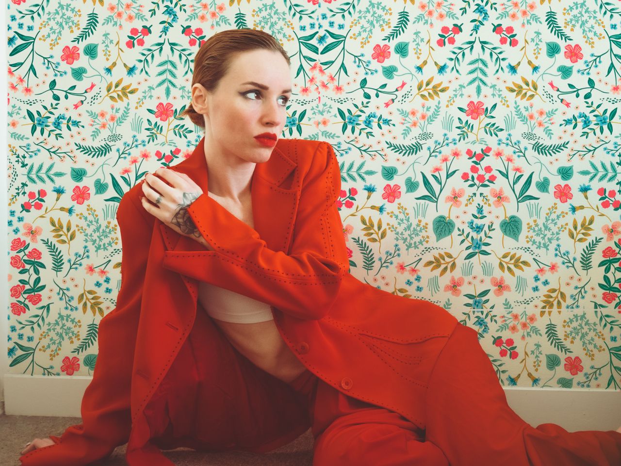 elegant woman in a red blazer sits against a vibrant floral wallpaper