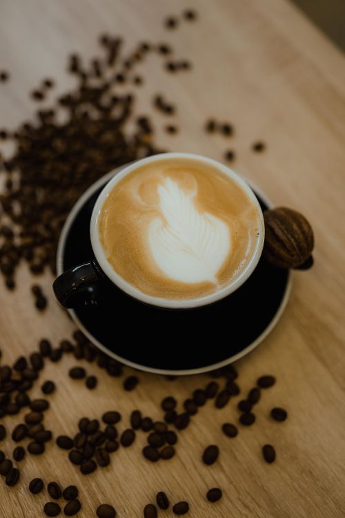 Free A warm cappuccino with elegant latte art served beside a macaron on a wooden table with scattered coffee beans. Stock Photo