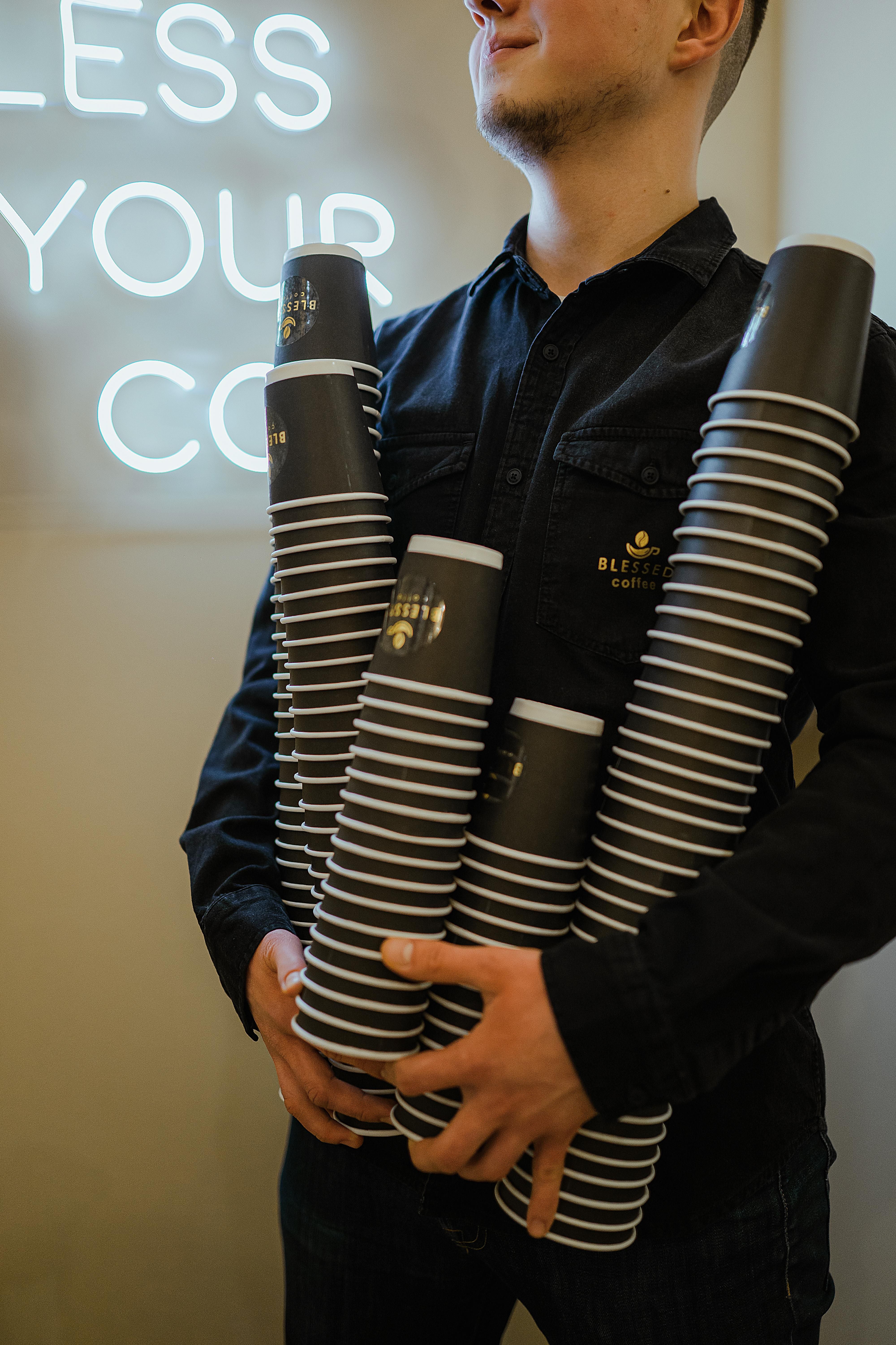Barista in dark uniform holding multiple stacks of coffee paper cups beneath neon sign indoors.