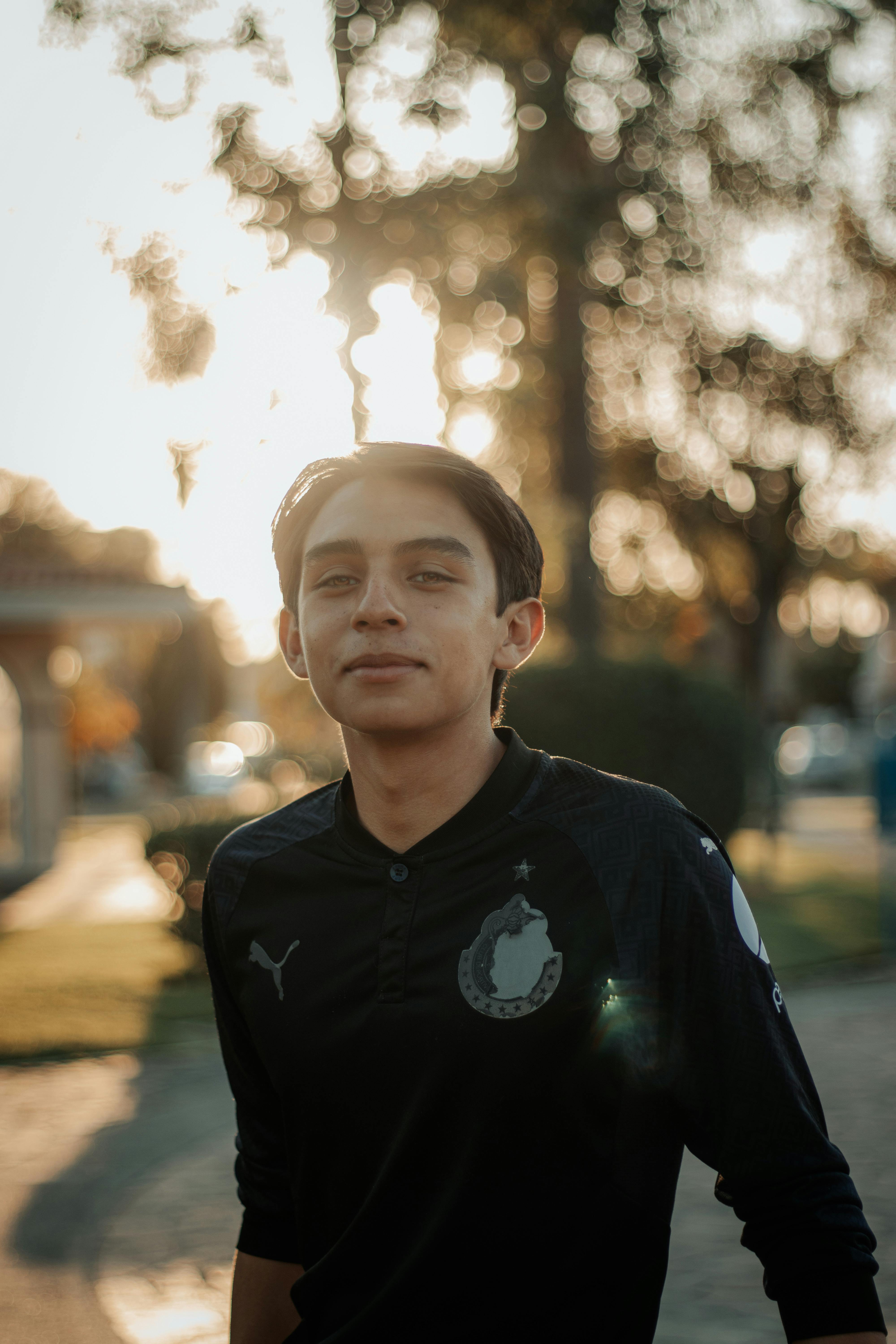 A young man in black stands with a slight smile, backlit by the sun in an outdoor setting.