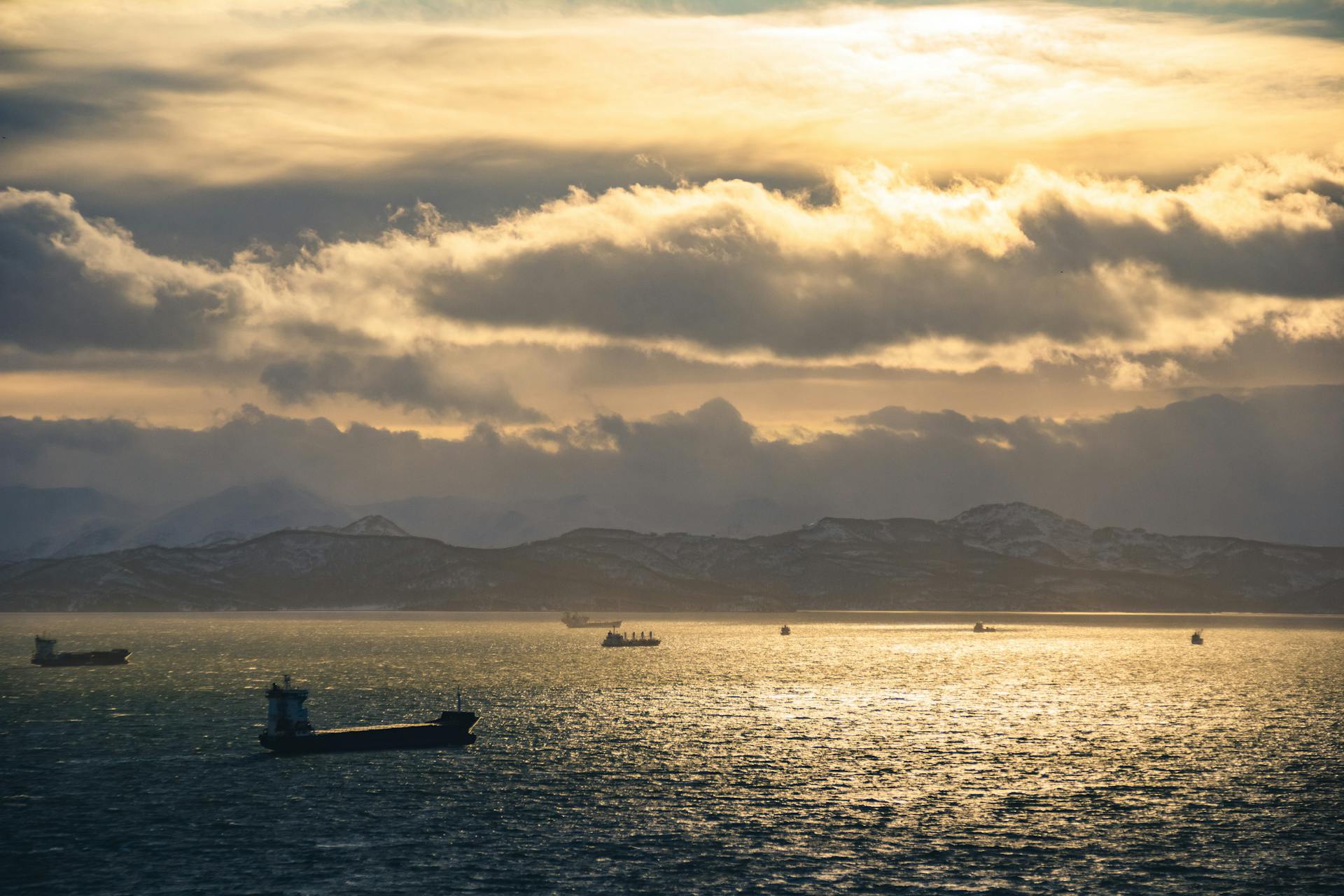 Golden sunset over a calm sea with silhouetted cargo ships and distant mountains.