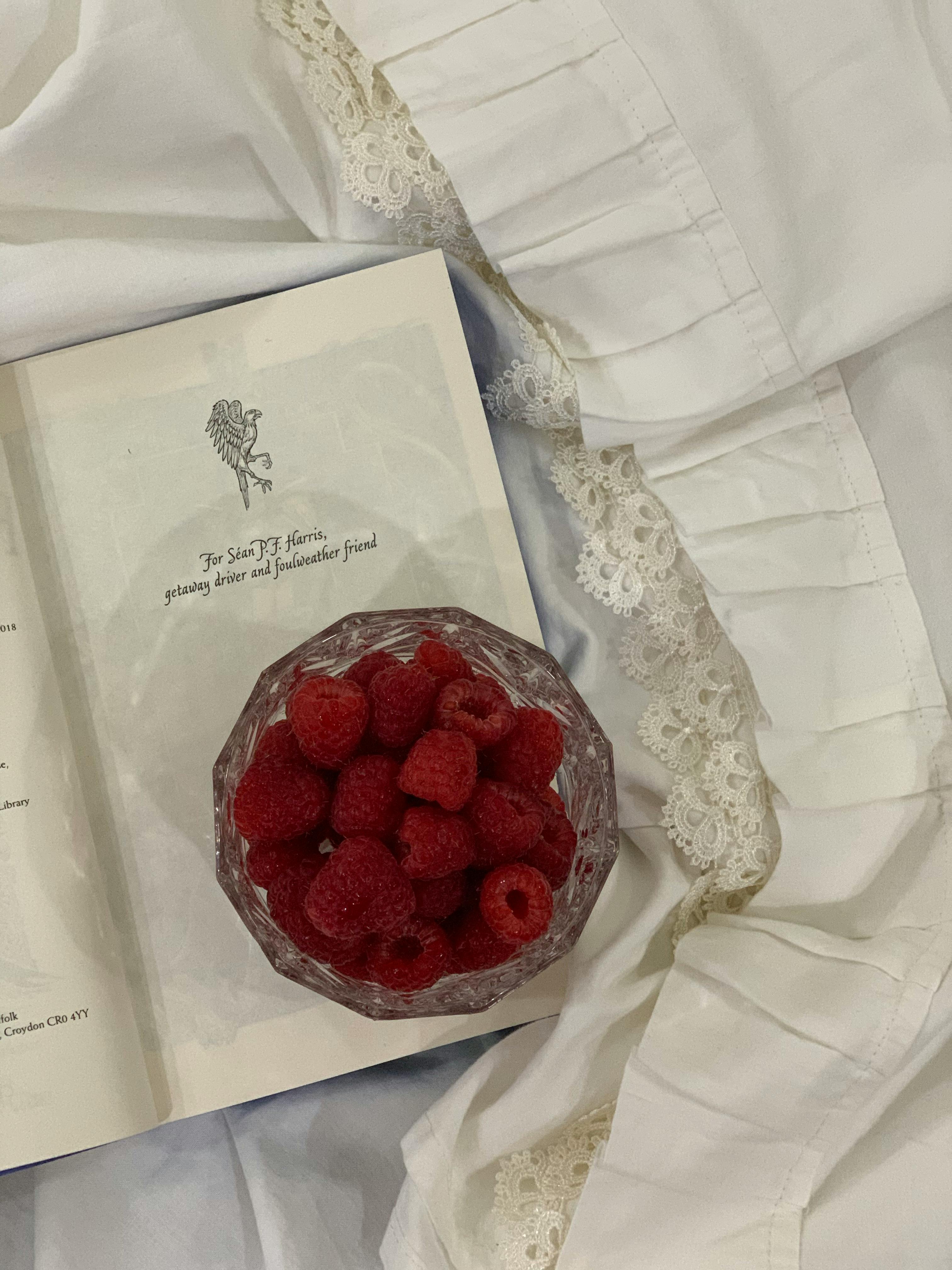 Flat lay of fresh raspberries in a glass bowl beside an open book on white fabric.