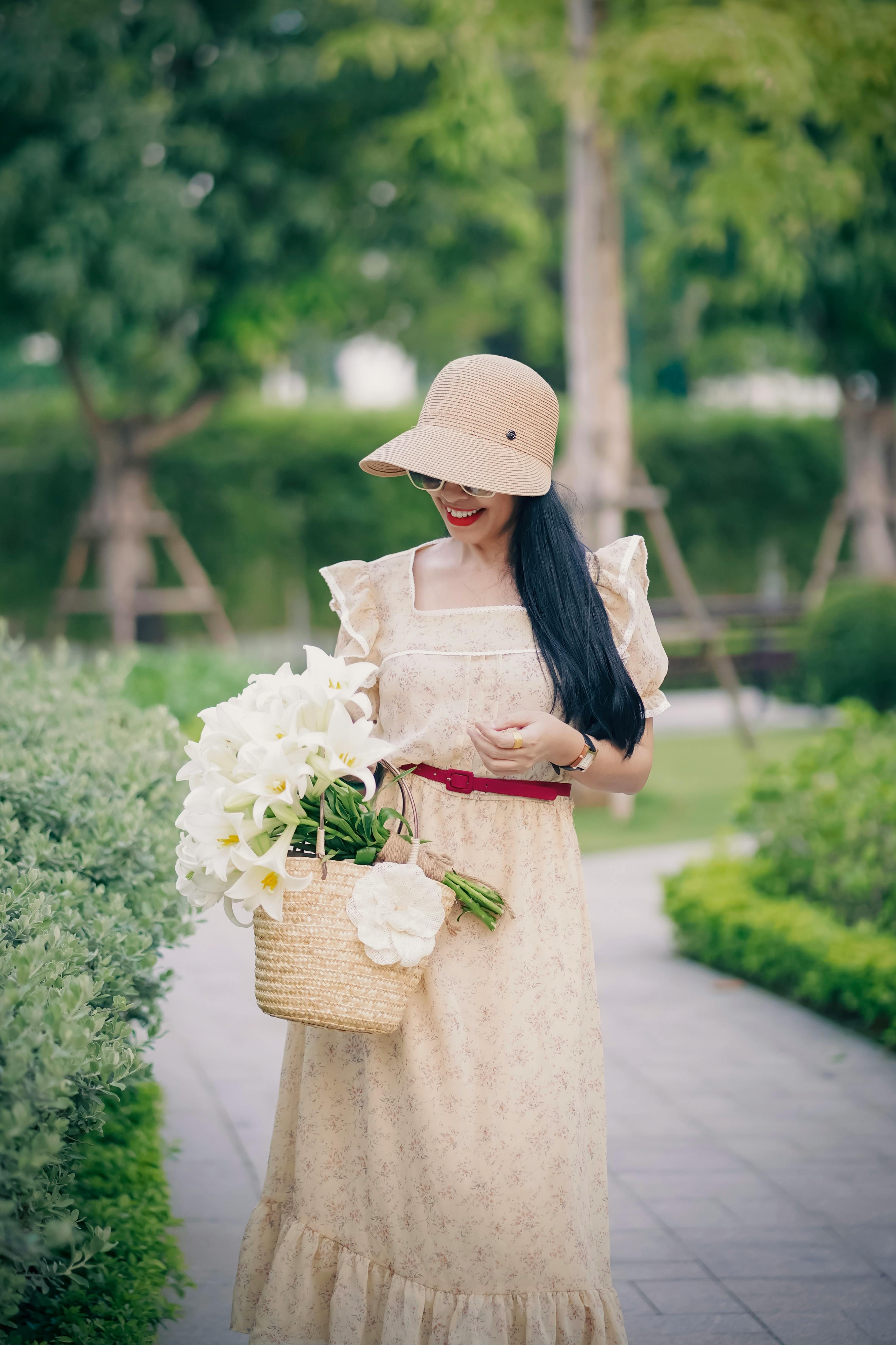 Fashionable woman in a public park wearing a beige dress and sun hat, holding a flower basket.
