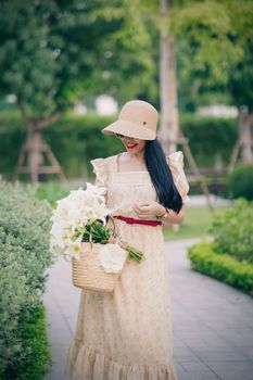 Fashionable woman in a public park wearing a beige dress and sun hat, holding a flower basket.