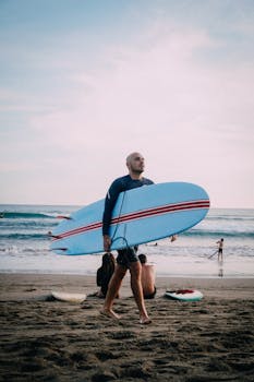 A man walks along the beach carrying a blue surfboard in a summer setting, perfect for water sports enthusiasts.