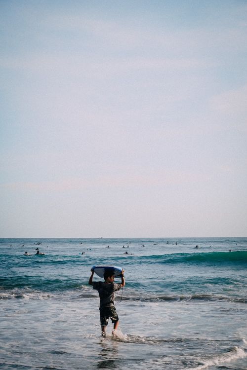 Free Young boy carrying surfboard at the beach with waves in the background, perfect for summer themes. Stock Photo