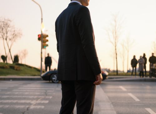 Free Man in a black suit crossing a city street at sunset, blending style with urban life. Stock Photo