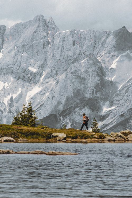 Free A lone hiker walks beside a serene lake with stunning snowcapped mountains in the background. Stock Photo
