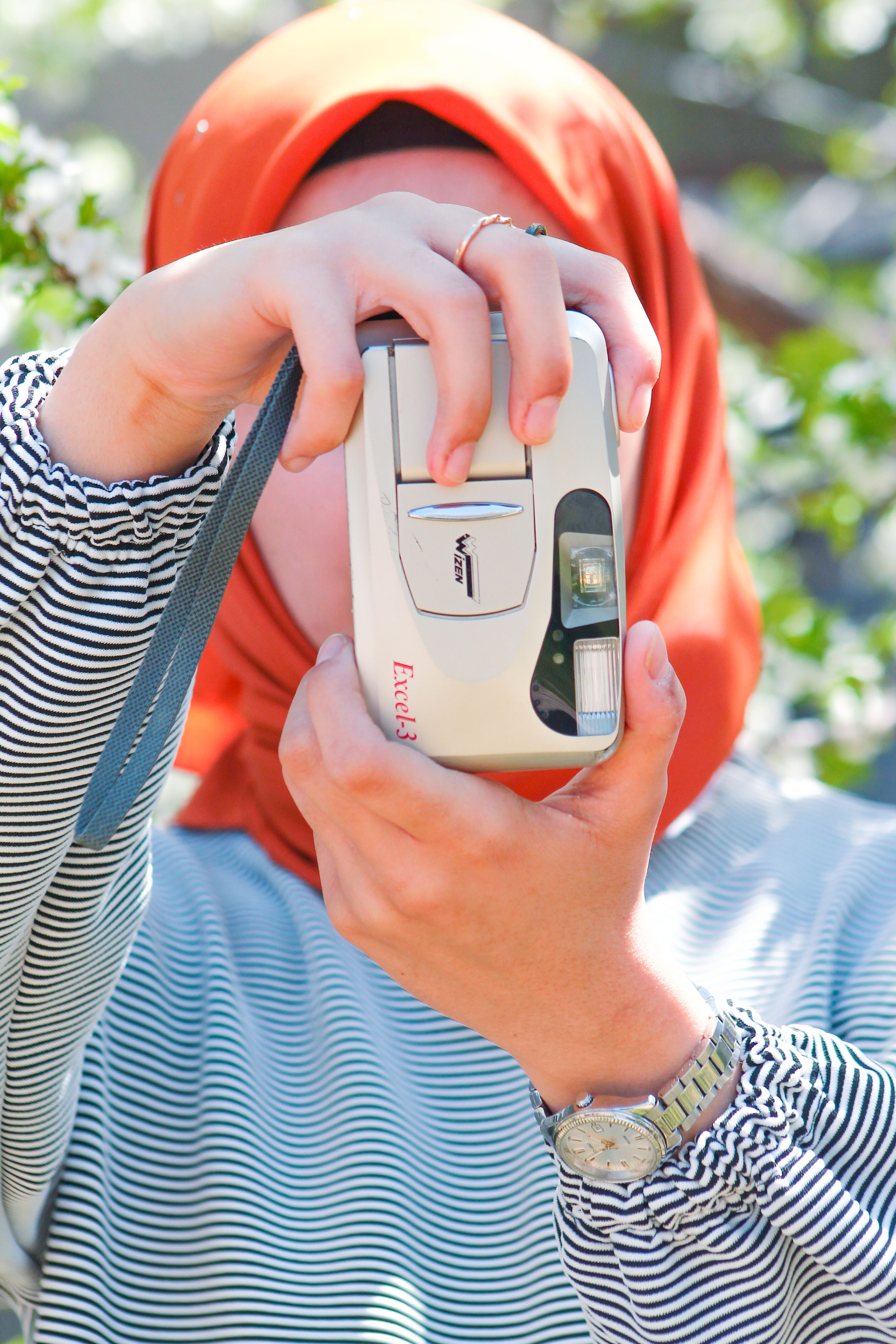 Woman in an orange hijab using a vintage point-and-shoot camera outdoors.