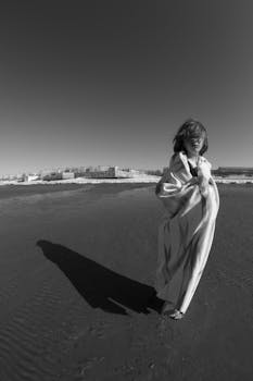 A lone woman wrapped in a blanket stands on a wide sandy beach, casting a long shadow.