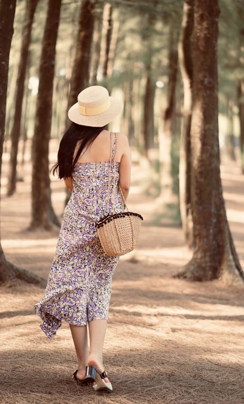 Free A woman in a straw hat and floral dress walking through a sunny pine forest path. Stock Photo