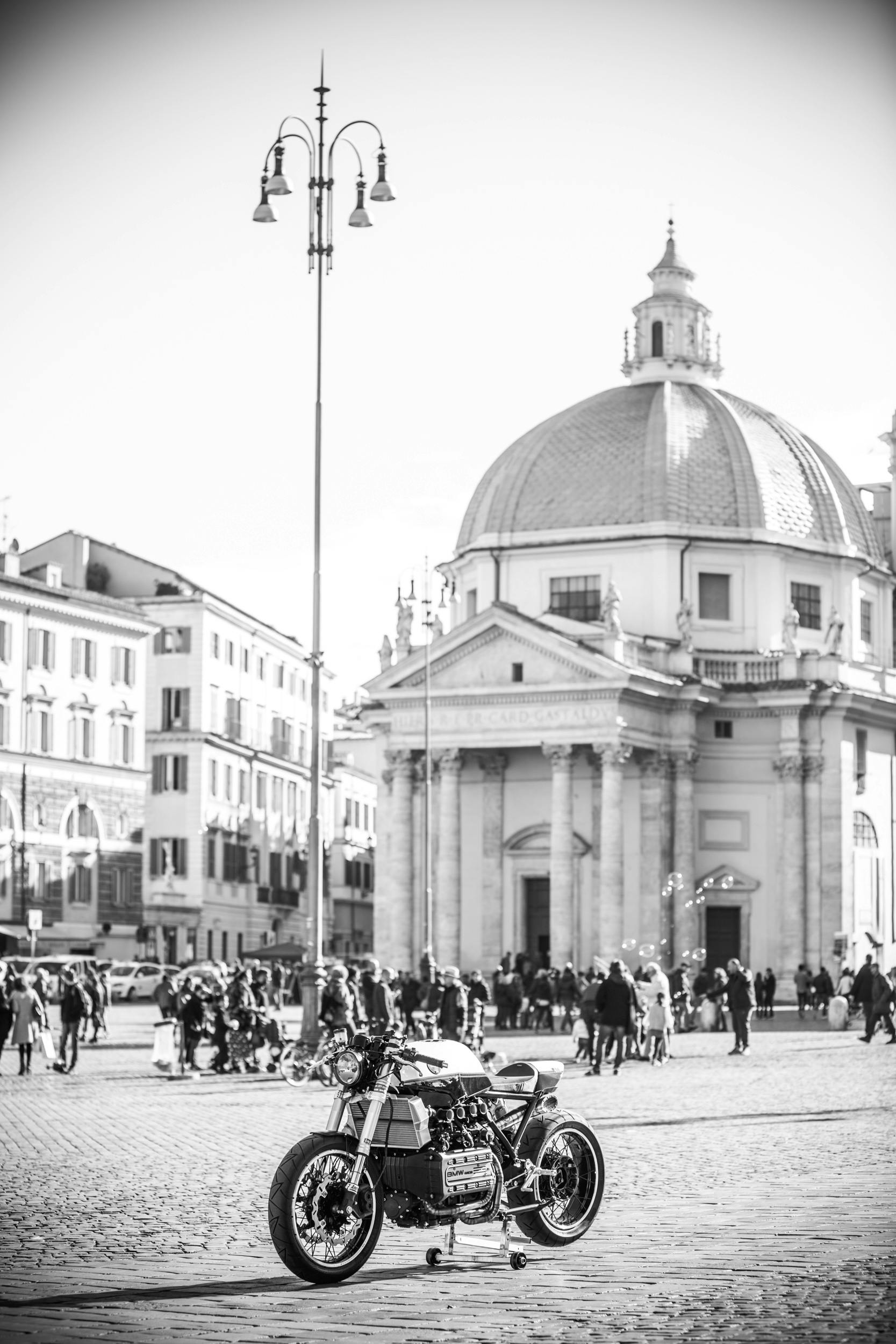 在罗马波波罗广场的黑白景观中，一辆老式摩托车穿梭于熙熙攘攘的人群之间