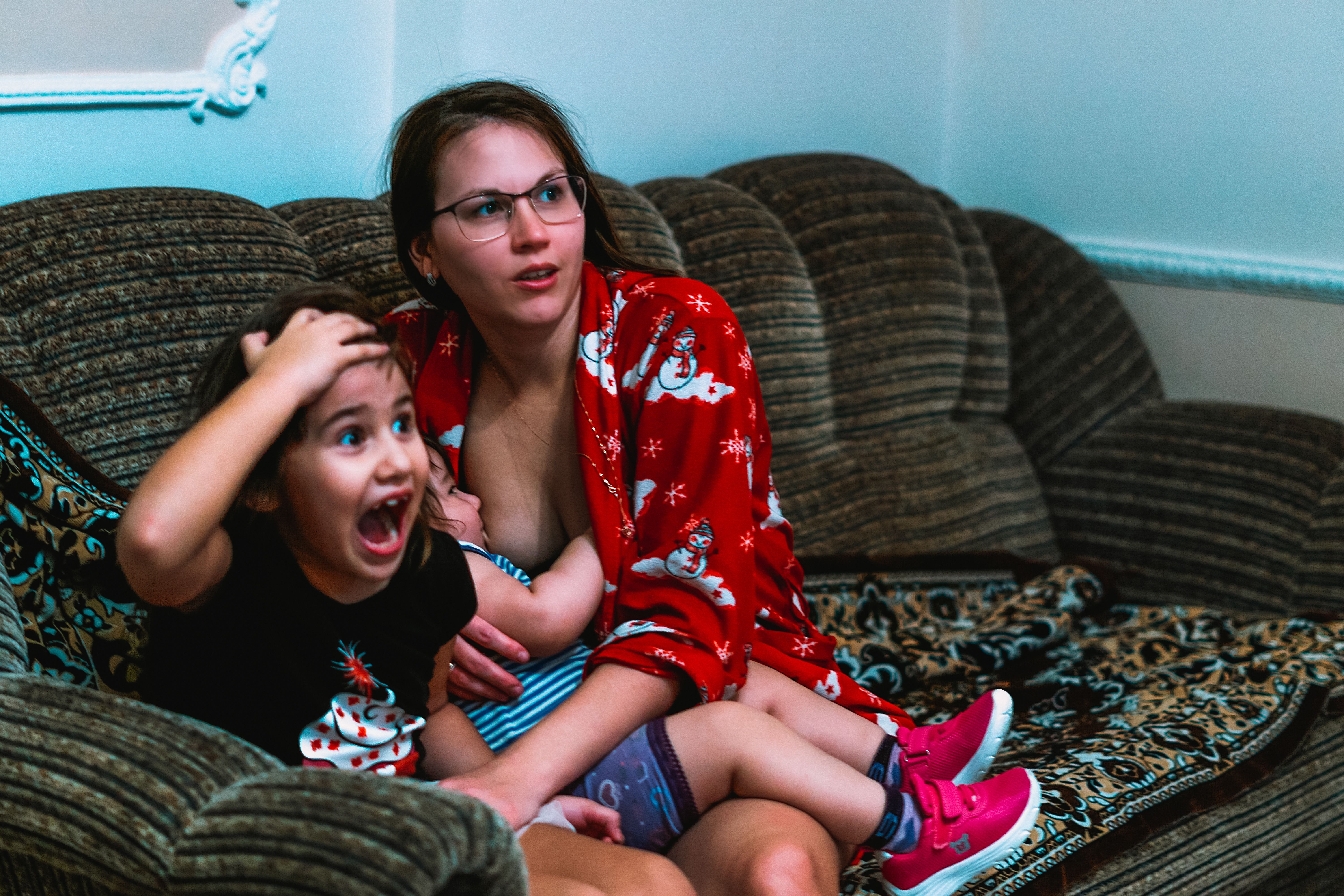 A mother and her children reacting with surprise while sitting on a sofa indoors.