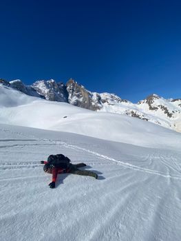 A backpacker enjoys a moment of freedom lying in the snow under a clear blue sky and majestic mountains.