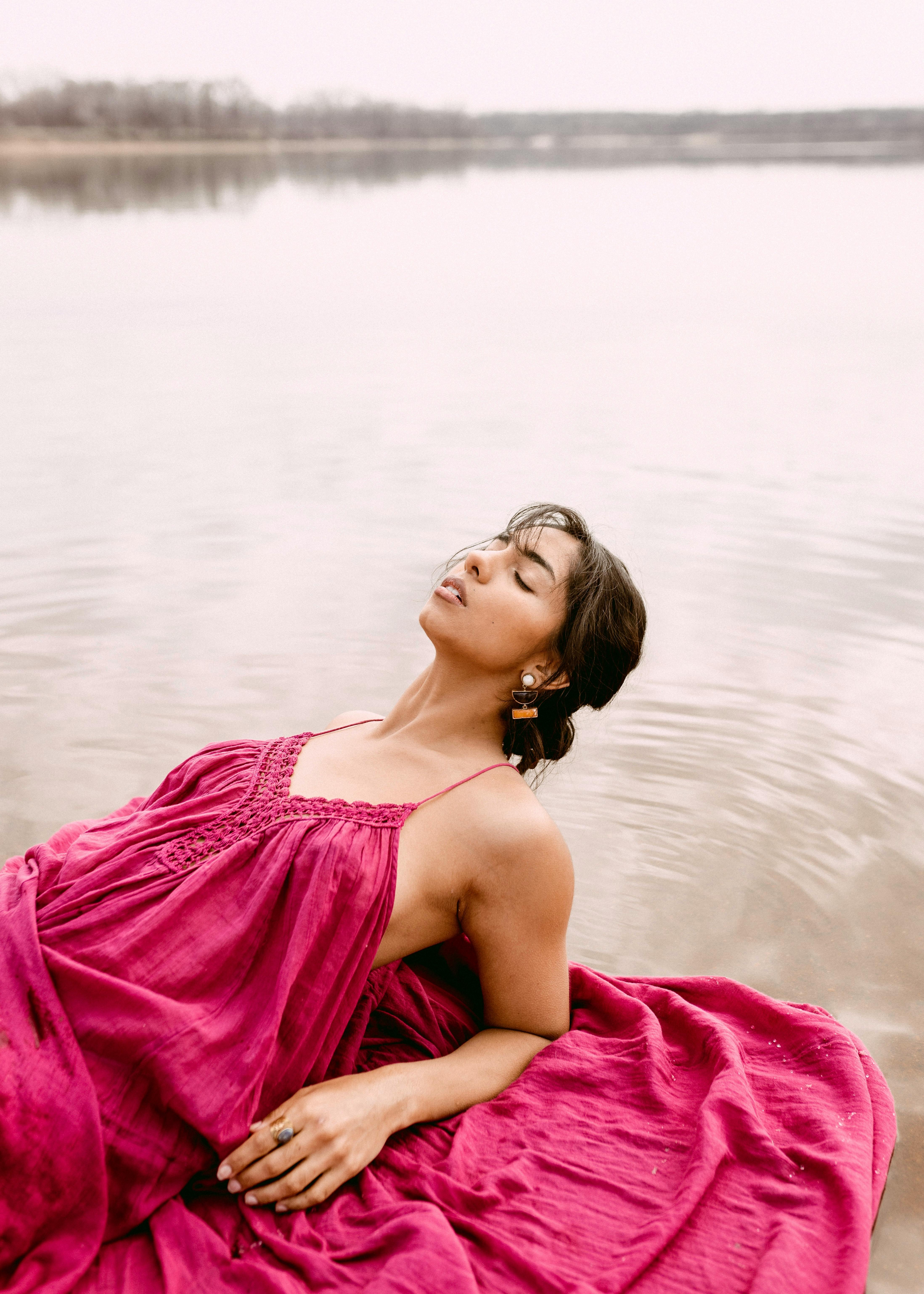 Woman in a red dress posing peacefully by a lake during the day.