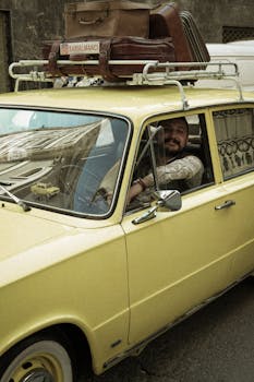 A smiling man sits in a vintage yellow car with luggage on the roof rack.