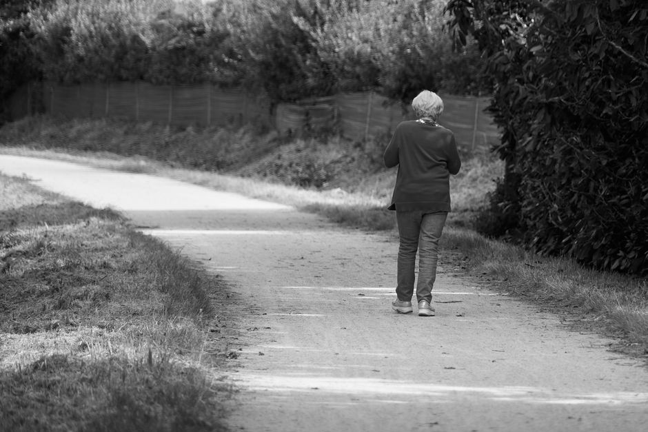 Black and white image of a senior woman walking down a quiet path, emphasizing solitude.