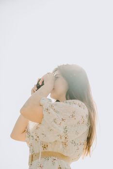 A young woman in a floral white dress taking a picture against a bright sky.