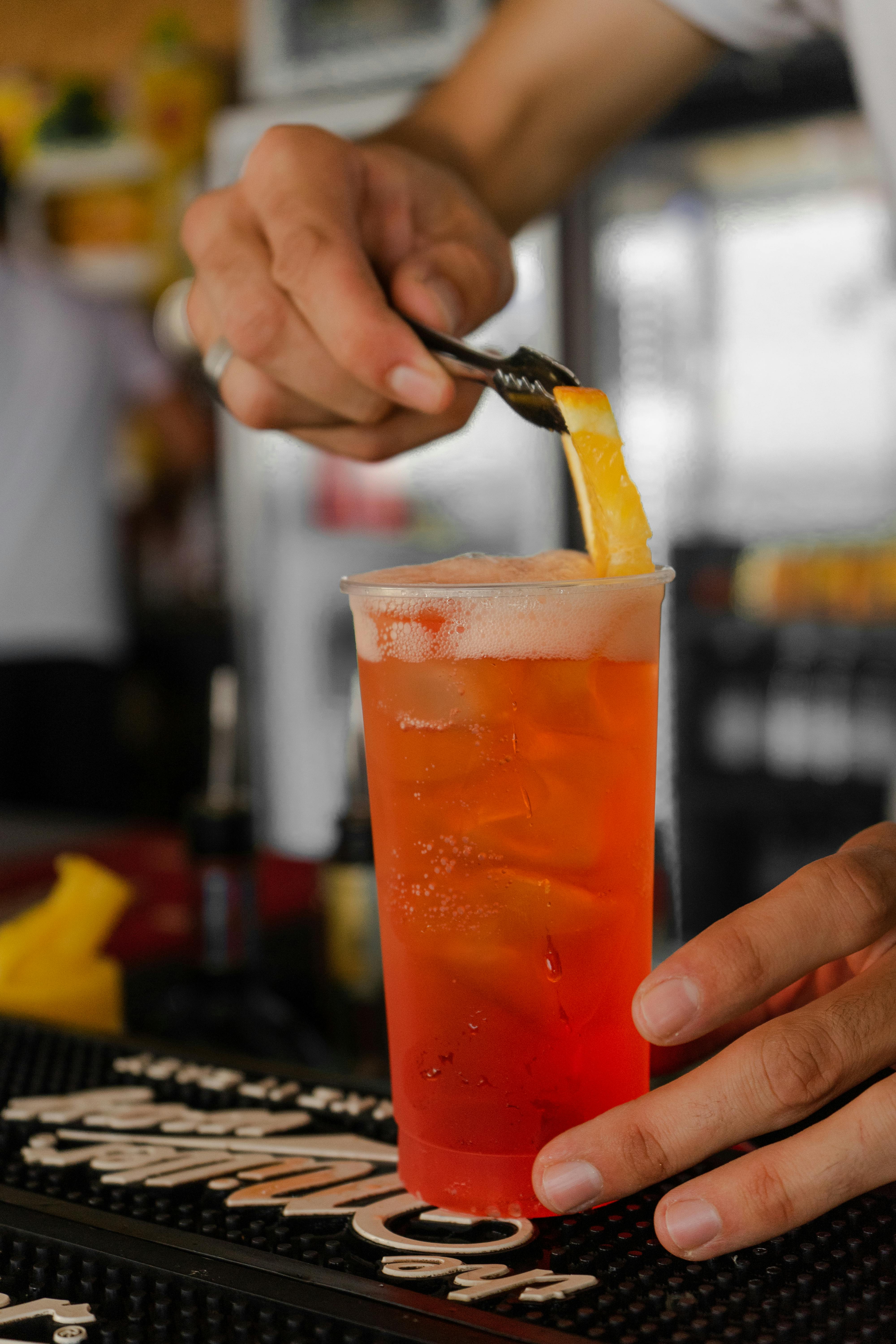 A bartender garnishing a vibrant orange cocktail with a slice of fruit using tongs, showcasing bar craftsmanship.