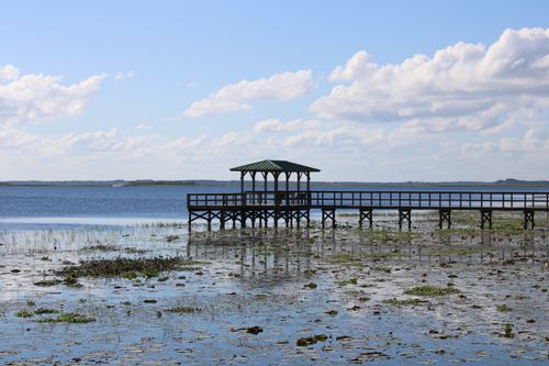 Free A picturesque wooden dock extends into a serene lake under a blue sky with fluffy clouds. Stock Photo