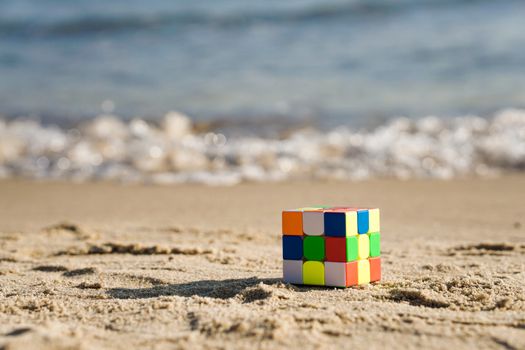 A colorful Rubik's cube placed on the sandy shore with ocean waves in the background.