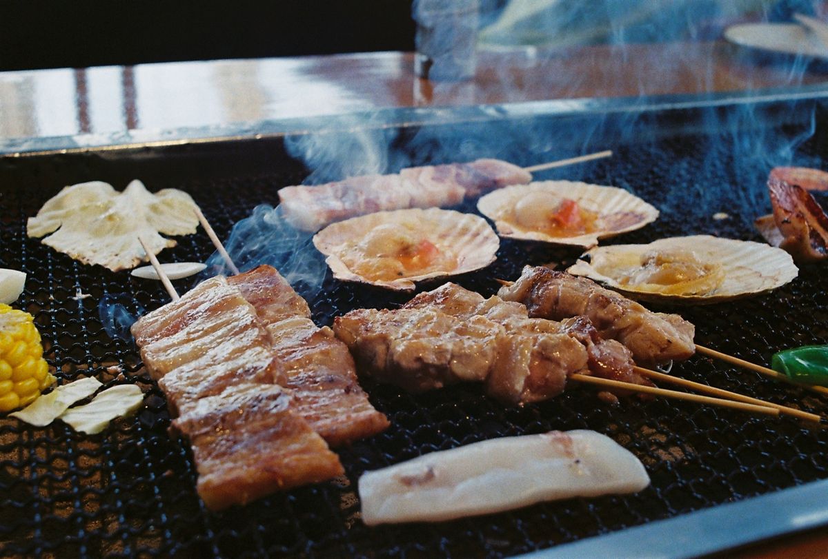 Grilled meat and scallop skewers sizzling over open charcoal, smoke rising past a row of corn cobs roasting alongside them.
