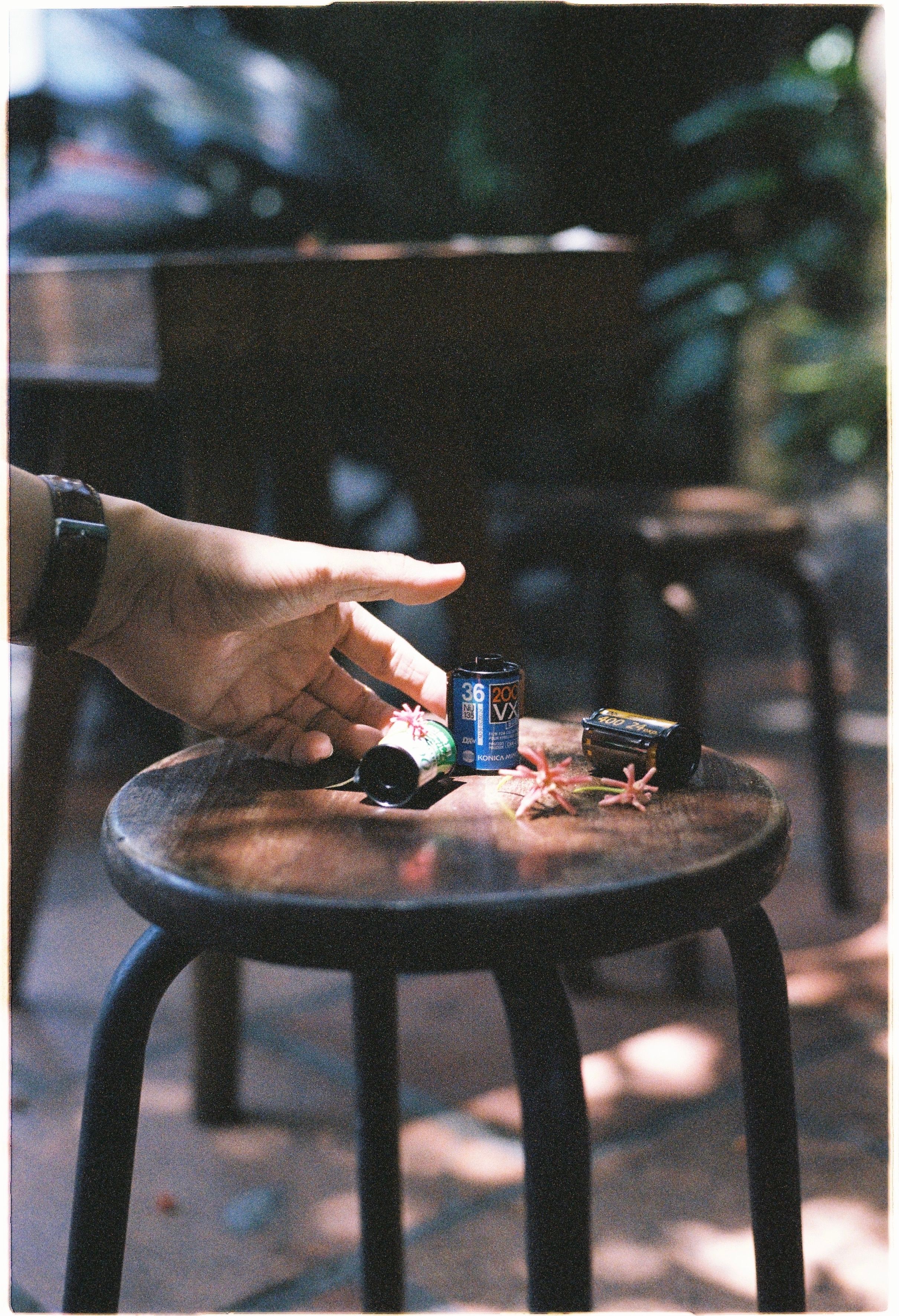 A hand reaching for film cartridges on a rustic wooden stool surrounded by small flowers.