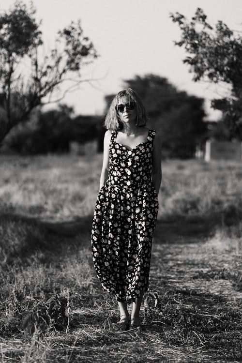 Grayscale Photo of a Woman in Floral Dress Standing on a Grassy Field