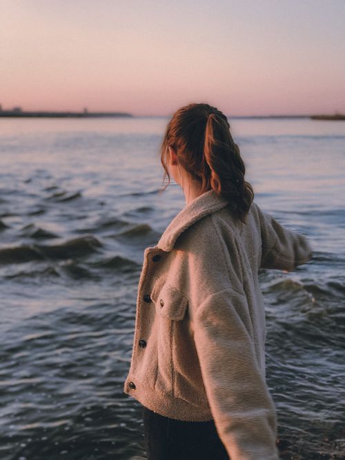 Free A woman stands by the seashore, enjoying the serene sunset. Her brown coat adds warmth to the scene. Stock Photo