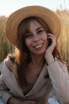 Charming woman in a straw hat and beige blazer smiling warmly in a sunny outdoor setting.