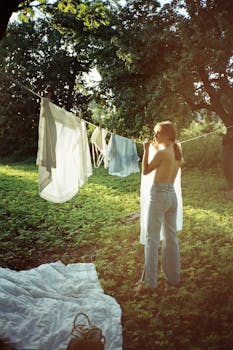 A woman hangs clothes on a line in a sunlit garden, evoking a sense of tranquility and rural life.