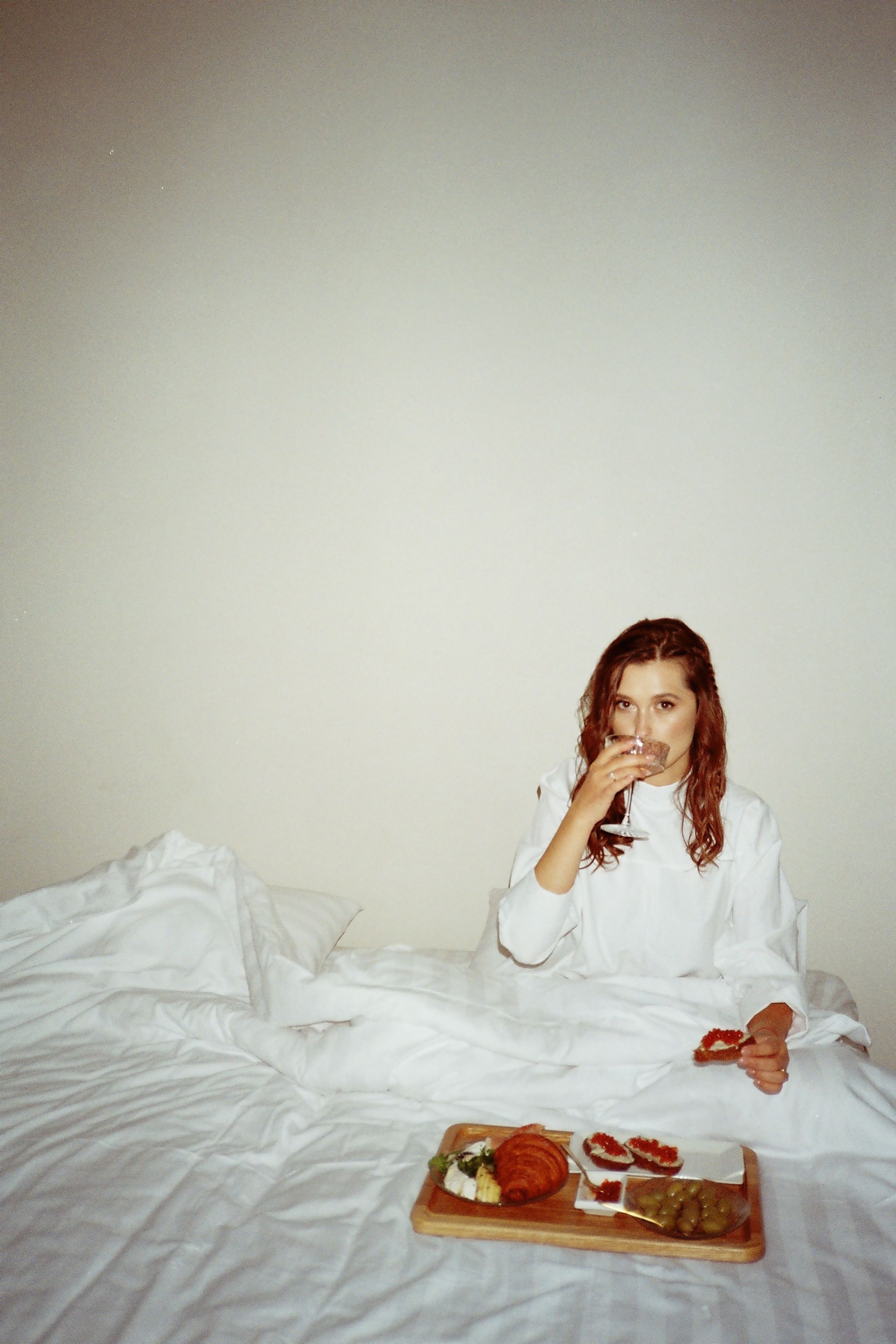 Relaxed woman sipping wine and enjoying breakfast in a serene bedroom setting.