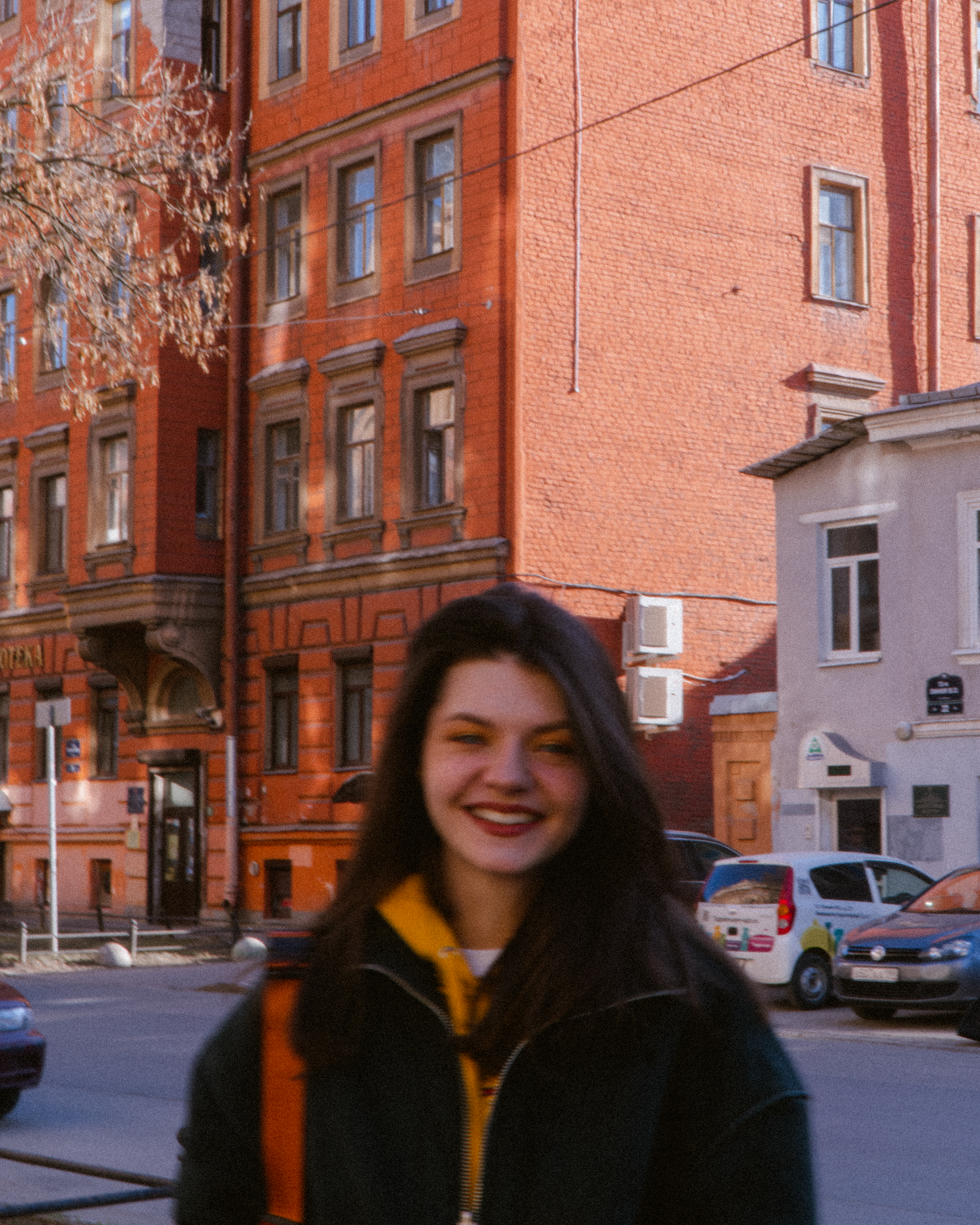 A smiling woman standing on a sunny street in Saint Petersburg, Russia, with red brick buildings behind her.