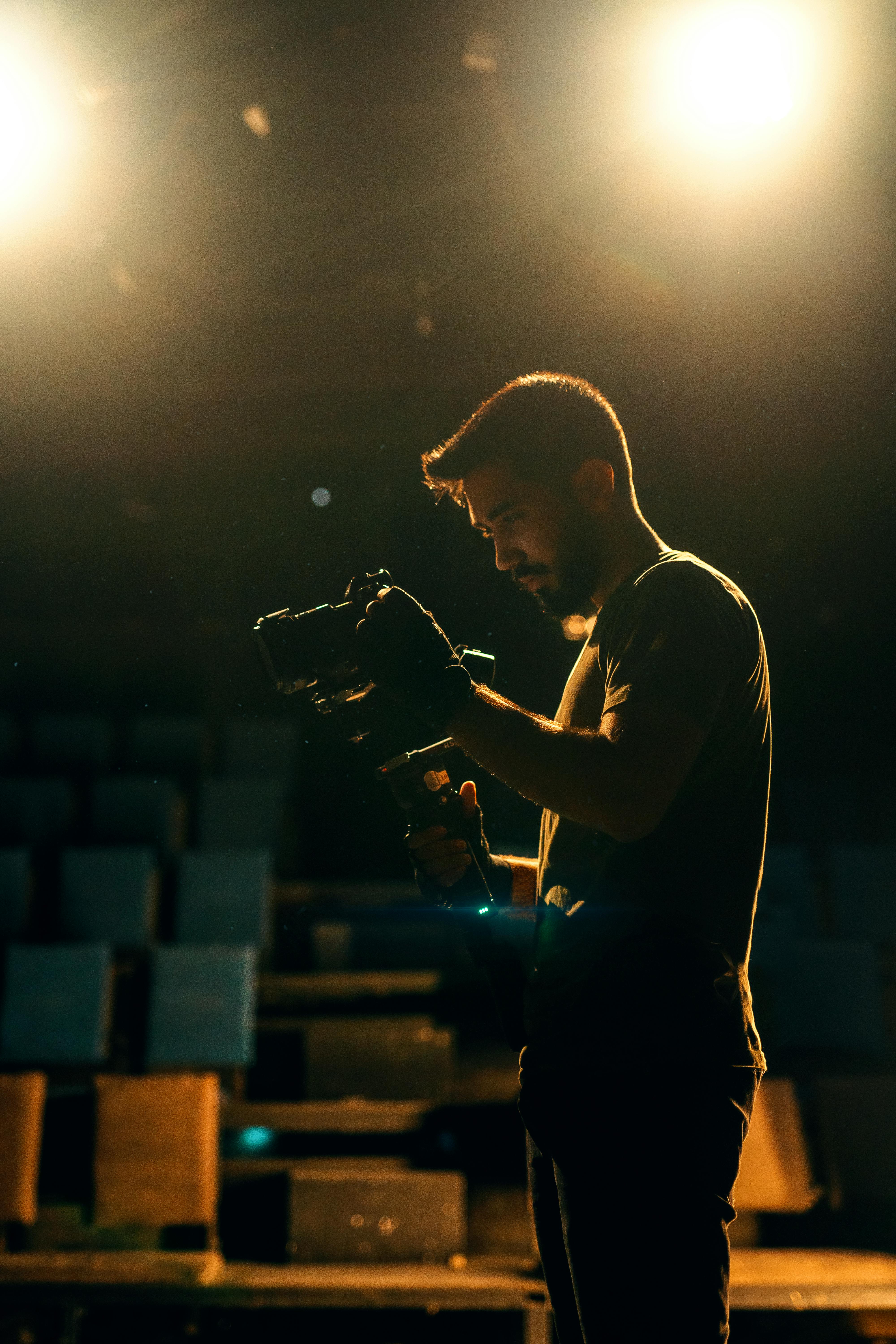 Free A filmmaker adjusts his camera under bright stage spotlights, creating a dramatic scene. Stock Photo