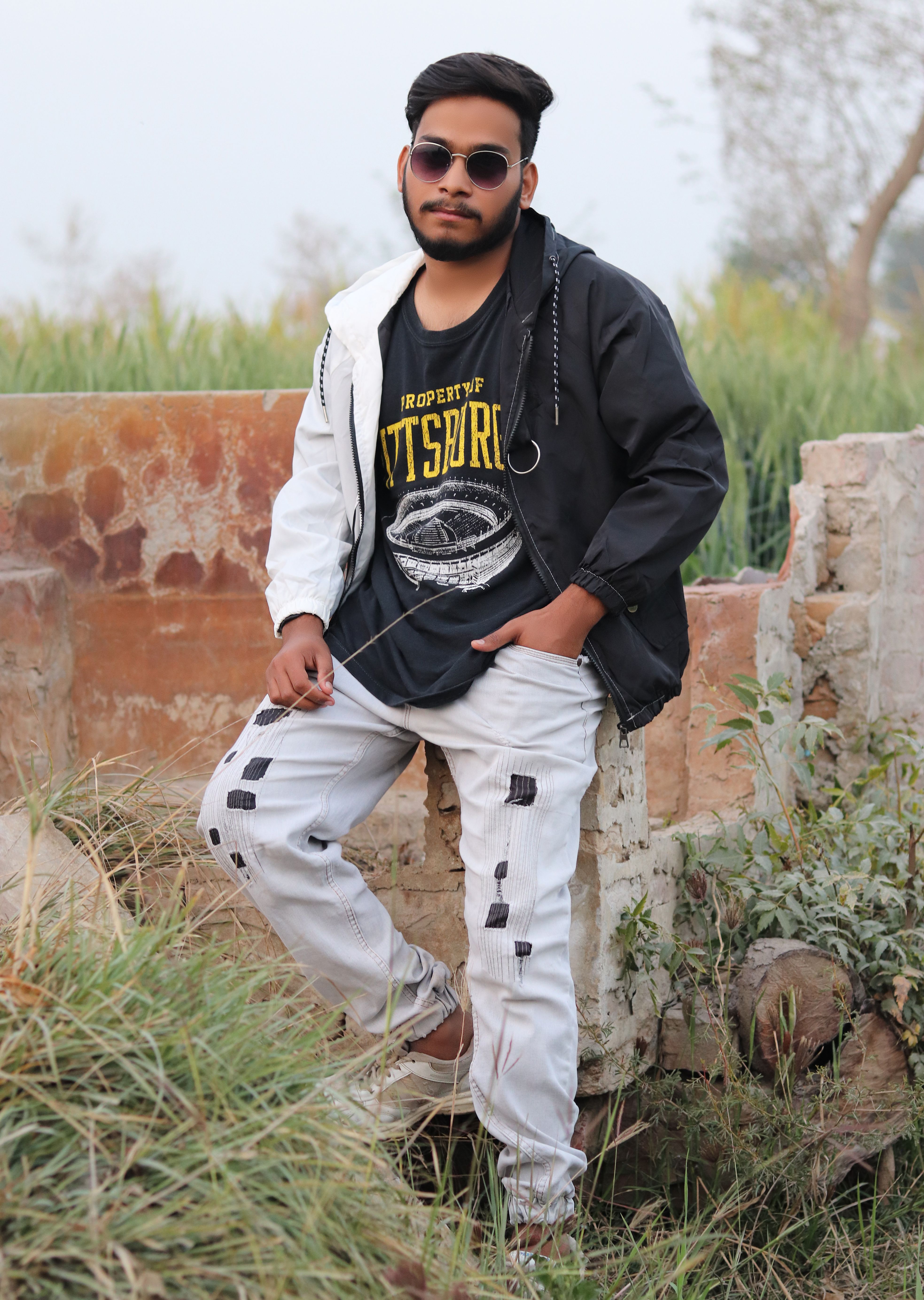 Outdoor photo of a stylish man sitting against a rustic brick wall, wearing a hoodie and sunglasses.