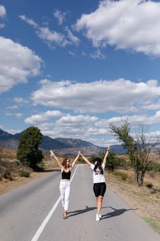 Two women joyfully walking with hands up on an open countryside road under a clear sky.