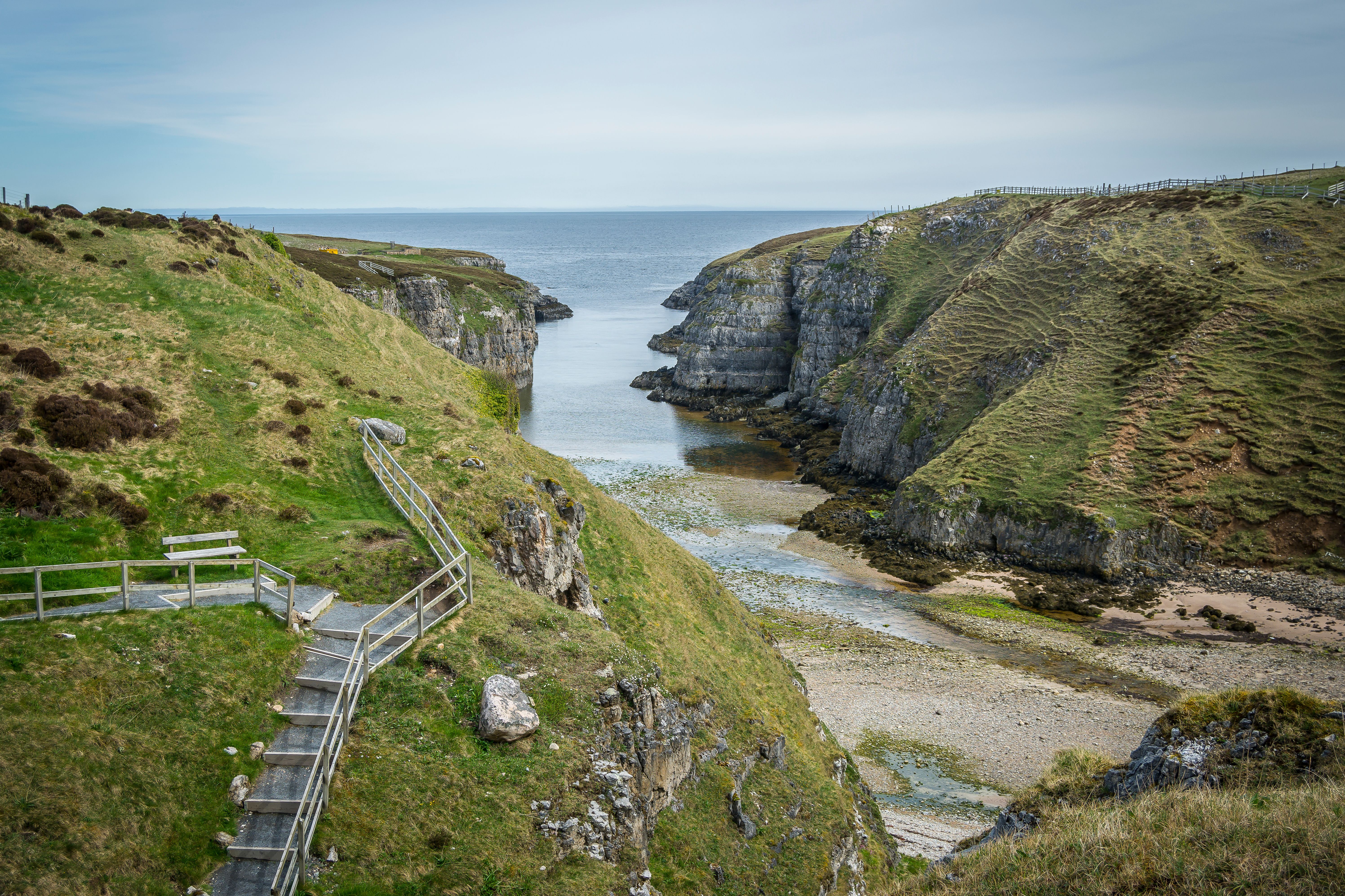 Scenic view of coastal cliffs and wooden path leading to calm sea under a clear sky.
