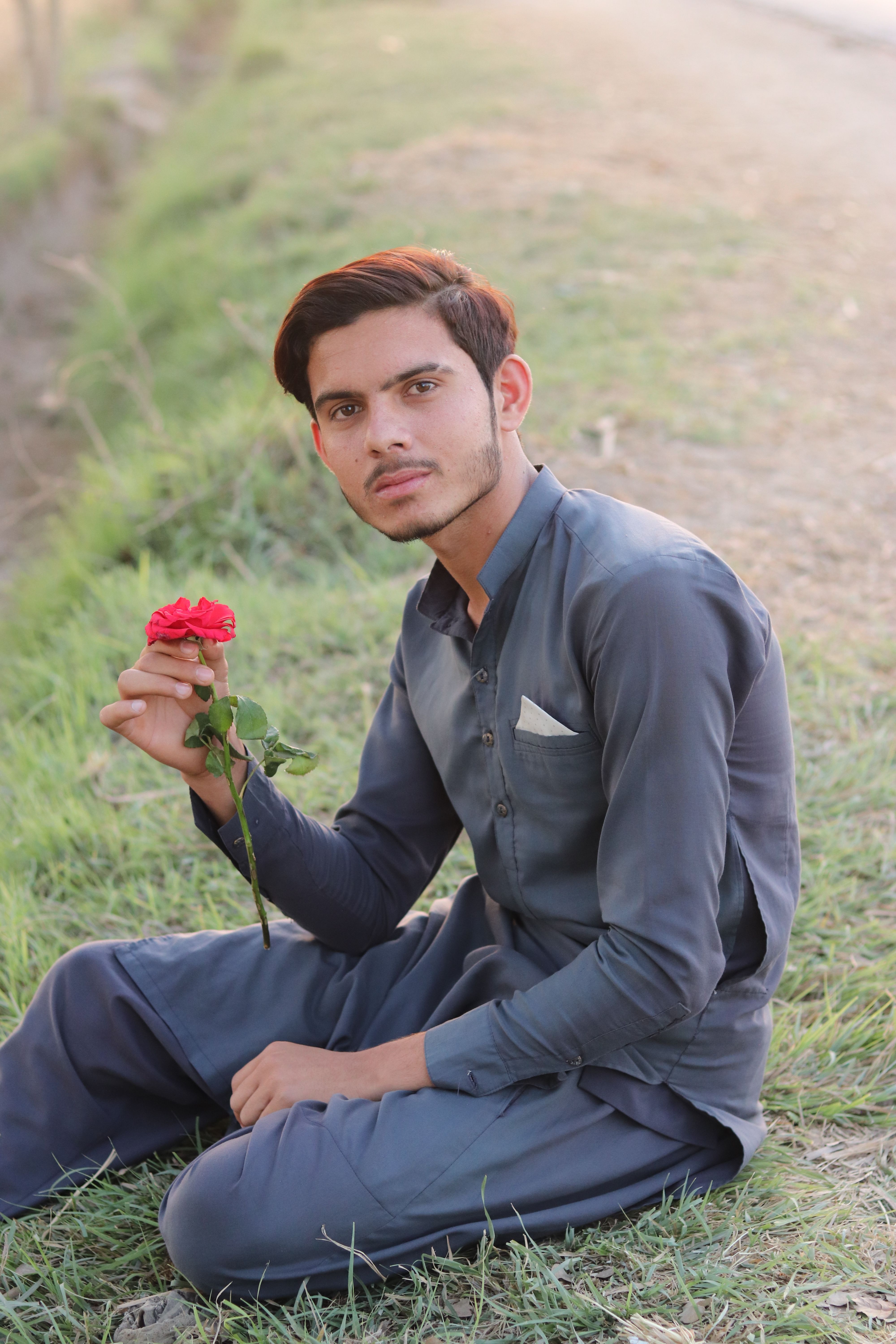 A man in blue attire sits on grass holding a red rose, conveying serenity.