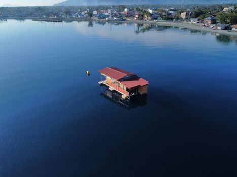 A serene aerial view of a red wooden floating house on a calm river near a town.