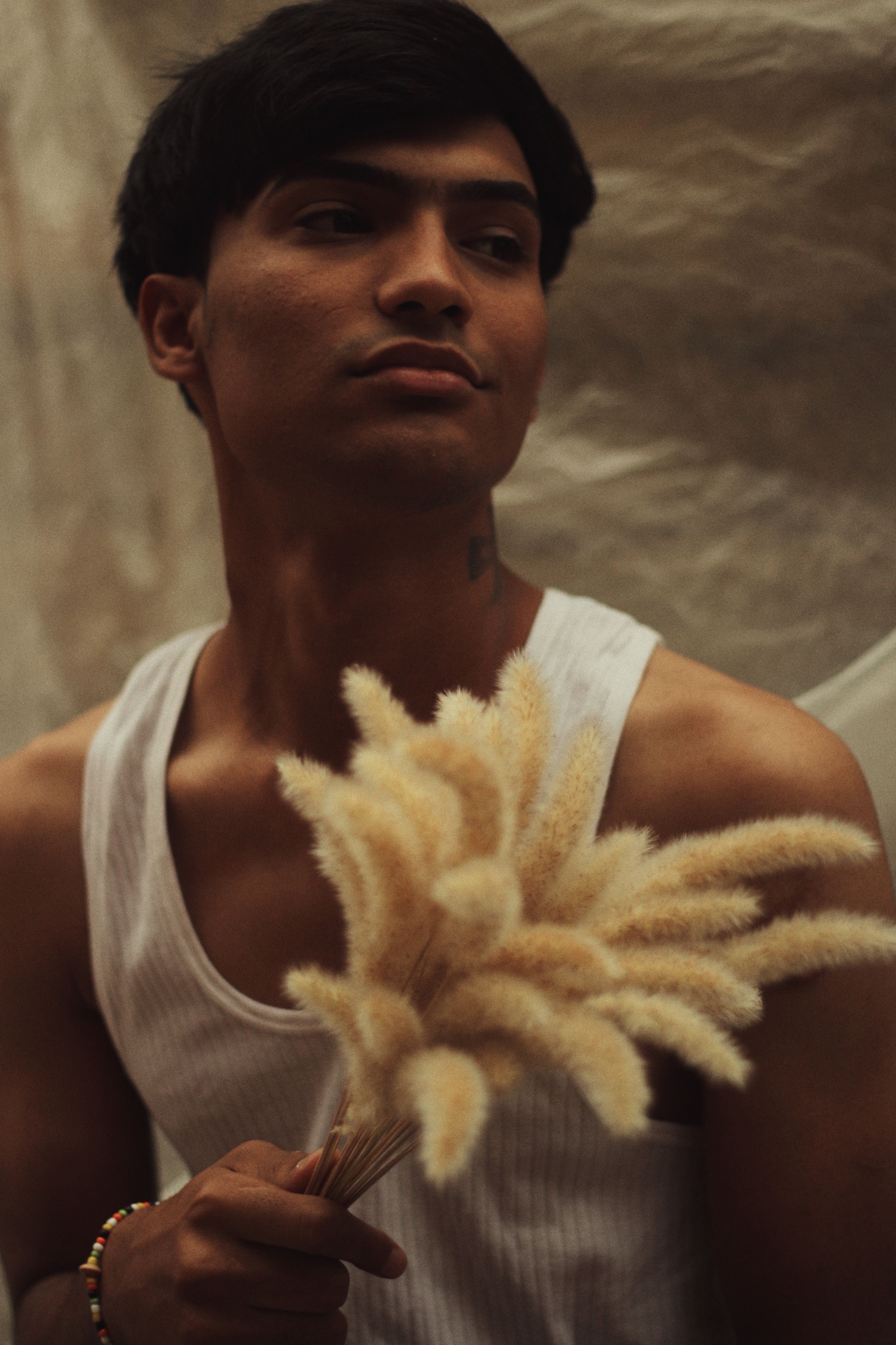 Young man in a white tank top posing indoors holding pampas grass.